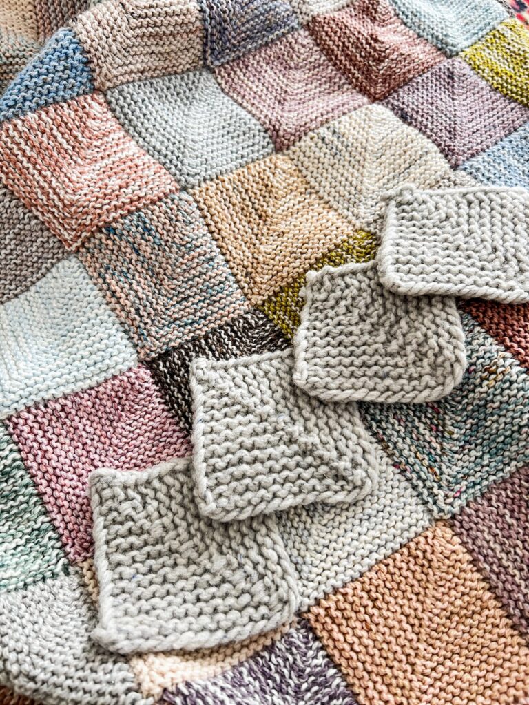 Four gray mitered squares are laid out in a diagonal row across a multicolored mitered square blanket.