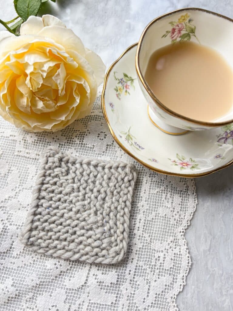 A gray mitered square formed with sk2togpsso decreases sits on a white crocheted doily next to a teacup full of milky tea and a yellow rose.