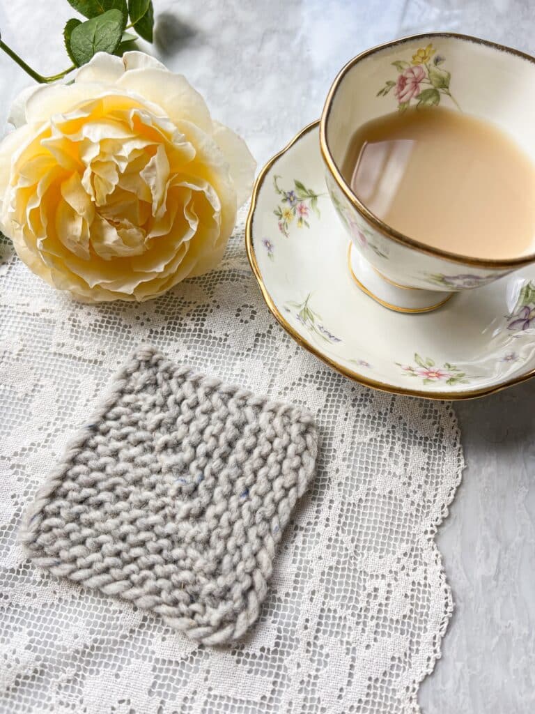 A gray mitered square formed with CDD decreases sits on a white crocheted doily next to a teacup full of milky tea and a yellow rose.
