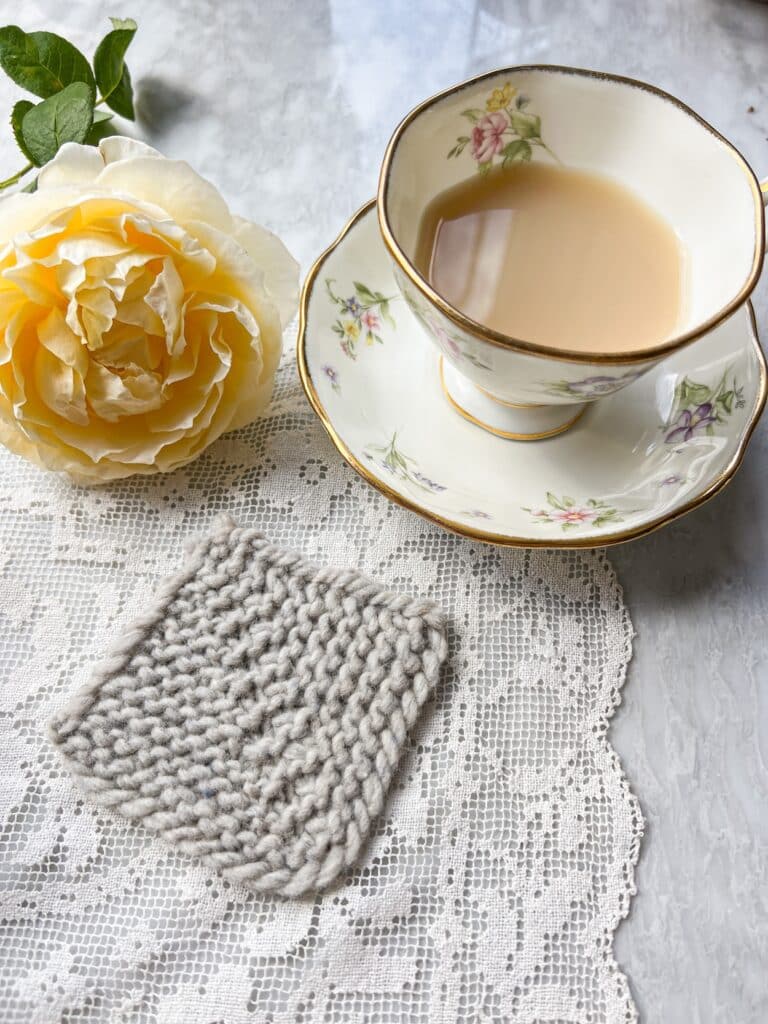 A gray mitered square formed with ssk, k1, k2tog decreases sits on a white crocheted doily next to a teacup full of milky tea and a yellow rose.