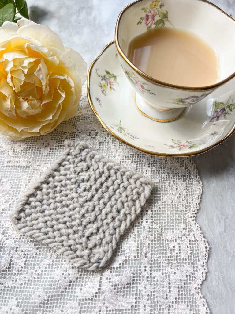 A gray mitered square formed with ssk, k2tog decreases sits on a white crocheted doily next to a teacup full of milky tea and a yellow rose.