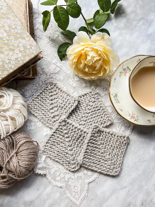 Four gray mitered squares sit on top of a lace doily next to a yellow rose, a teacup full of milky tea, some antique books, and two neutral-colored balls of yarn.