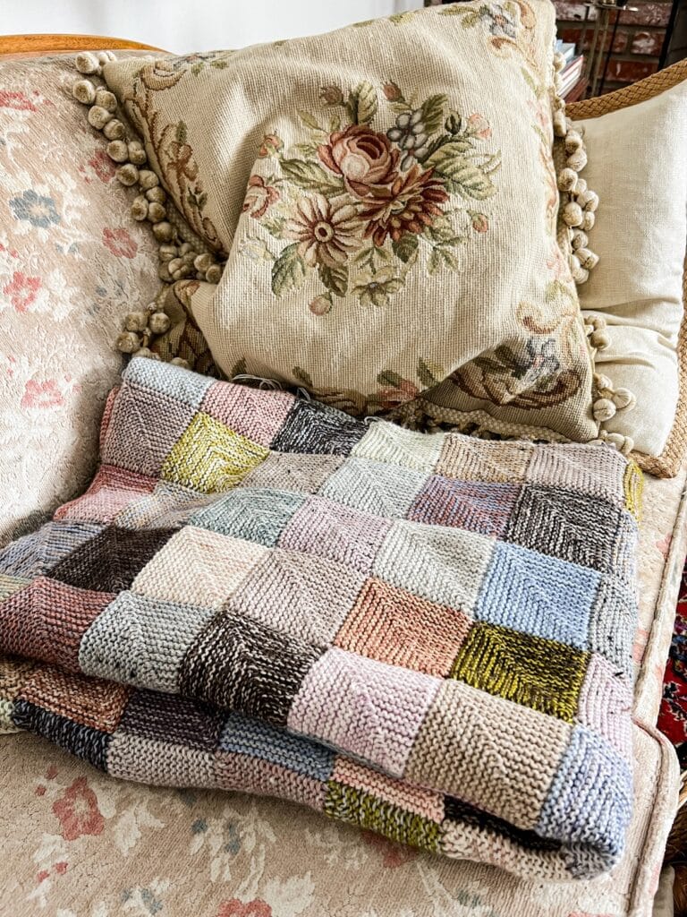 A multi-colored mitered square blanket is folded on a vintage sofa with faded floral upholstery. A needlepoint cushion sits next to the blanket.