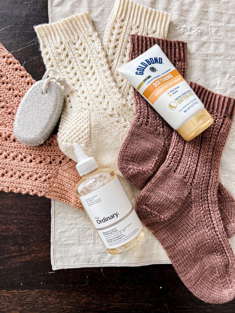 A top-down photo of three pairs of handknit socks in shades of pink, white, and brown. Interspersed among the socks are a pumice stone, a bottle of glycolic acid toner, and a tube of moisturizing foot cream.