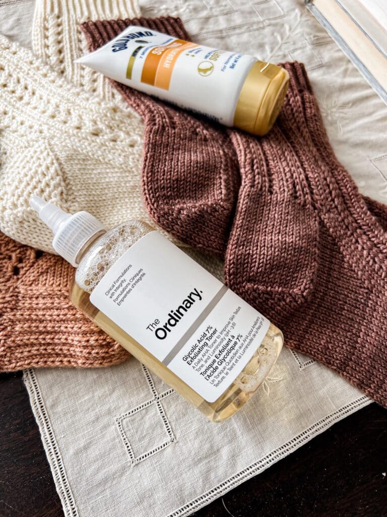 A close-up on a bottle of glycolic acid toner sitting on a small pile of pink, white, and brown handknit socks.
