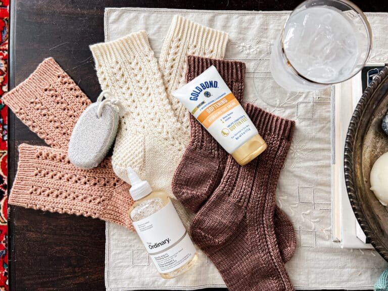 A top-down photo of three pairs of handknit socks in shades of pink, white, and brown. Interspersed among the socks are a pumice stone, a bottle of glycolic acid toner, a glass of ice water, and a tube of moisturizing foot cream.