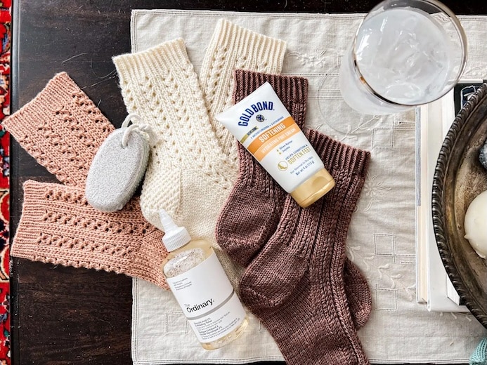 A top-down photo of three pairs of handknit socks in shades of pink, white, and brown. Interspersed among the socks are a pumice stone, a bottle of glycolic acid toner, a glass of ice water, and a tube of moisturizing foot cream.