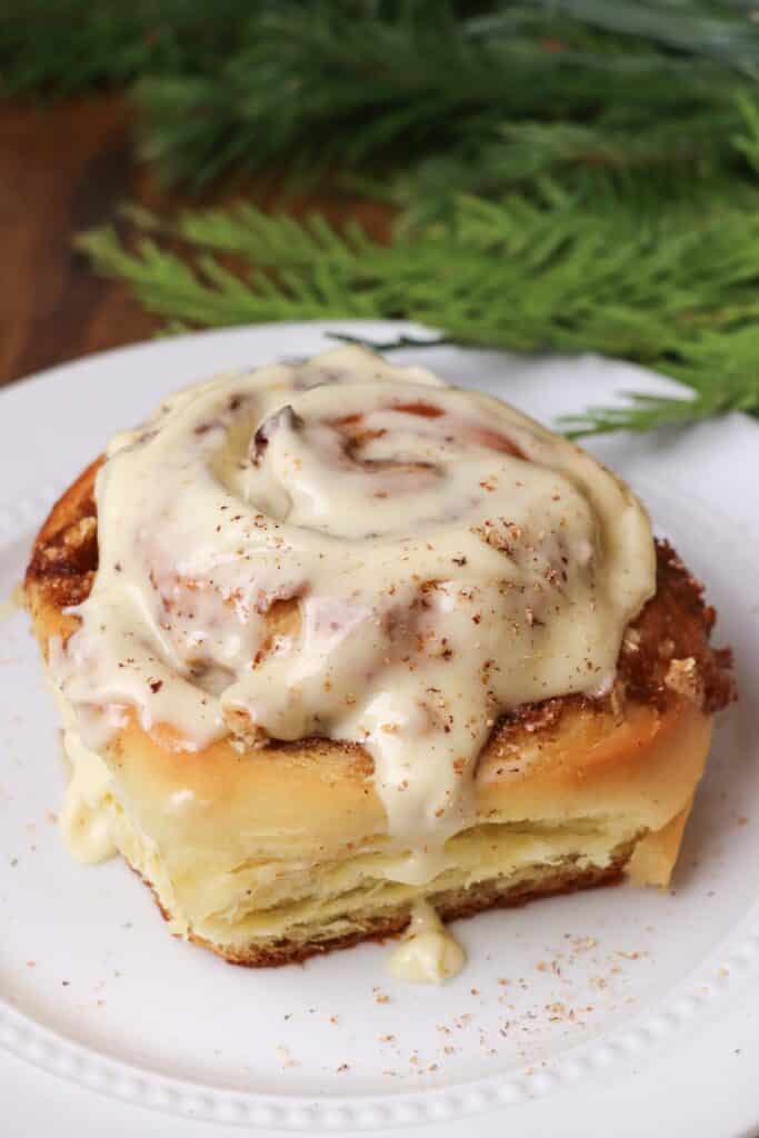 A cinnamon roll with a heaping dollop of cream cheese frosting sits on a white plate in front of a Christmas tree branch.