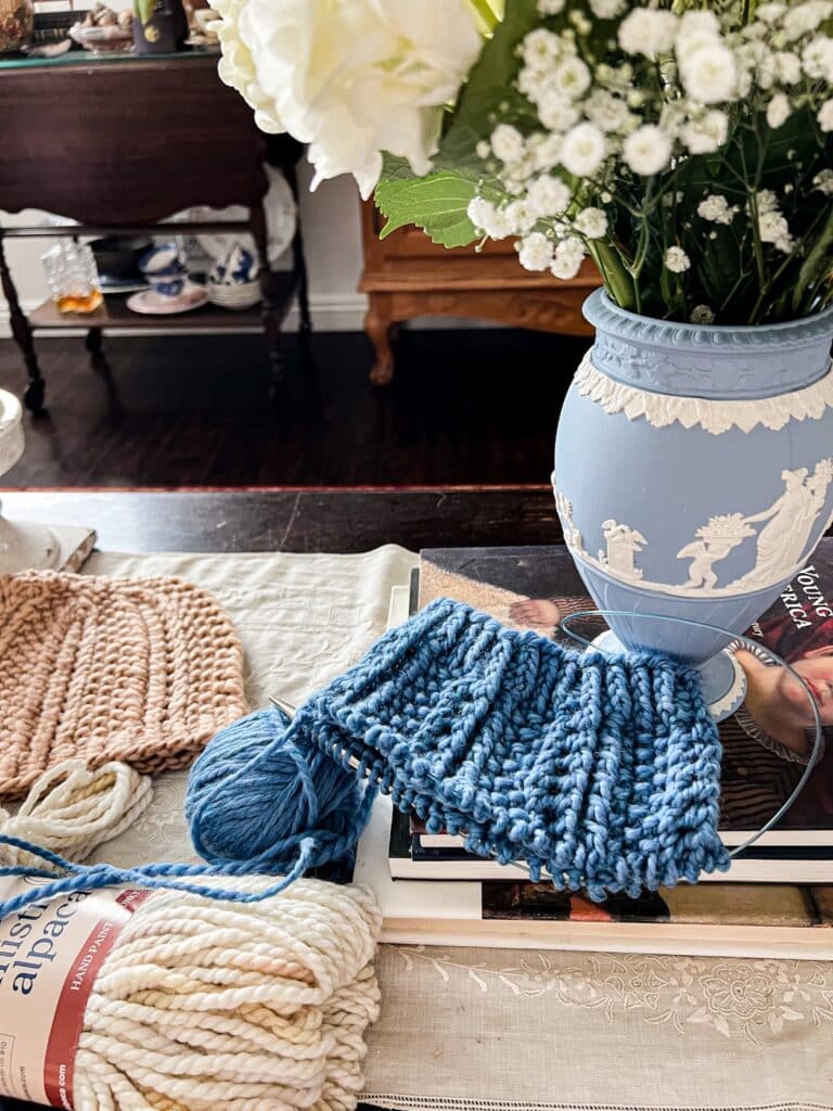 A blue chunky knit hat in progress sits on a small pile of books next to a light blue vase, a hank of creamy yarn, and a finished knit hat in a shade of dusty pink.