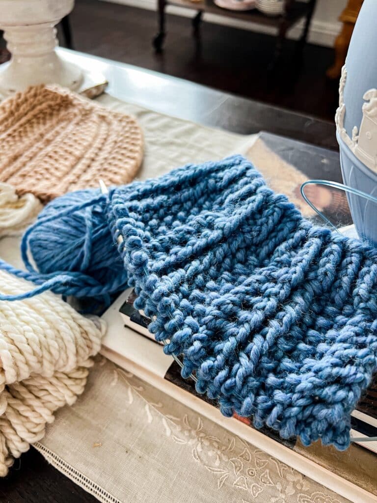 A close-up on a blue hat in progress. The hat is knit with chunky yarn and has lots of texture. It's sitting on a small pile of books next to a light blue vase.