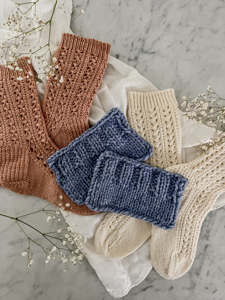 Two blue knit swatches with ribbing at the top sit on a crumpled white cloth surrounded by a pair of pink socks, a pair of white socks, and a few clumps of baby's breath flowers. The left blue swatch is bound off using the standard bind-off, while the other is bound off in pattern.