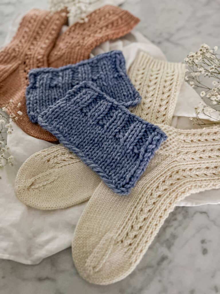 A low-angle photograph of two blue knit swatches surrounded by white and pink socks and a few sprigs of baby's breath. The top, foremost swatch is in focus and shows a ribbed edge bound off in pattern.