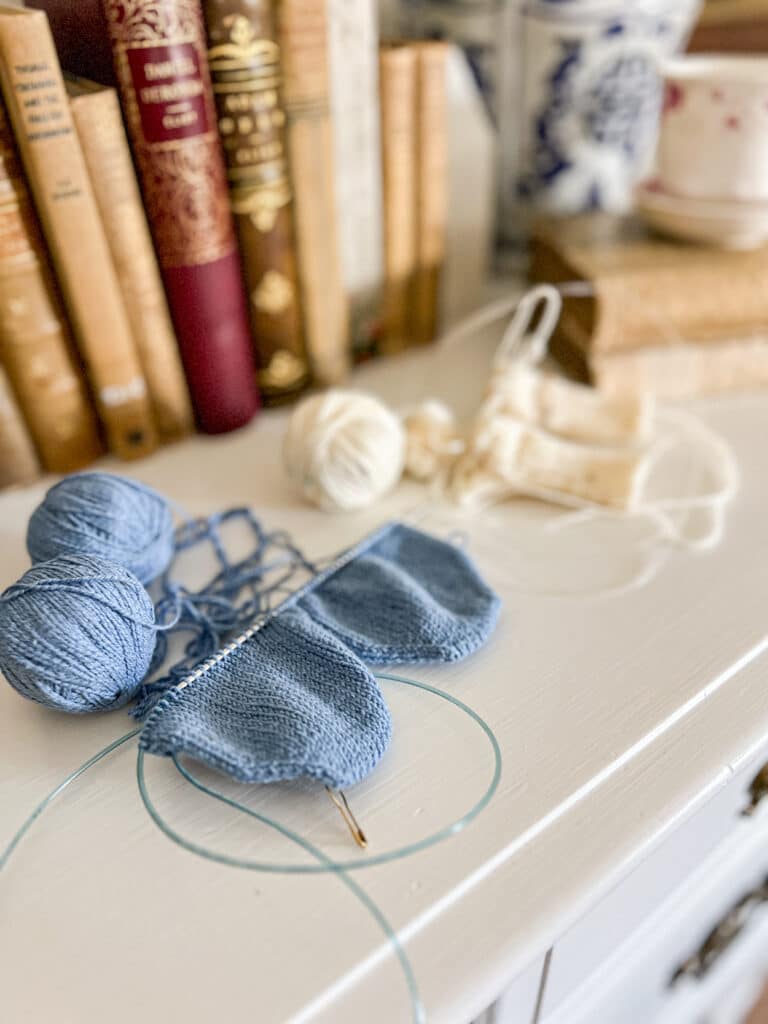 A side-angle photo showing a pair of blue adult knit socks in progress on a long circular needle and, blurred in the background, a pair of white baby socks in progress on a long circular needle. The blue socks are knit from the toes up while the white socks are knit from the cuff down.