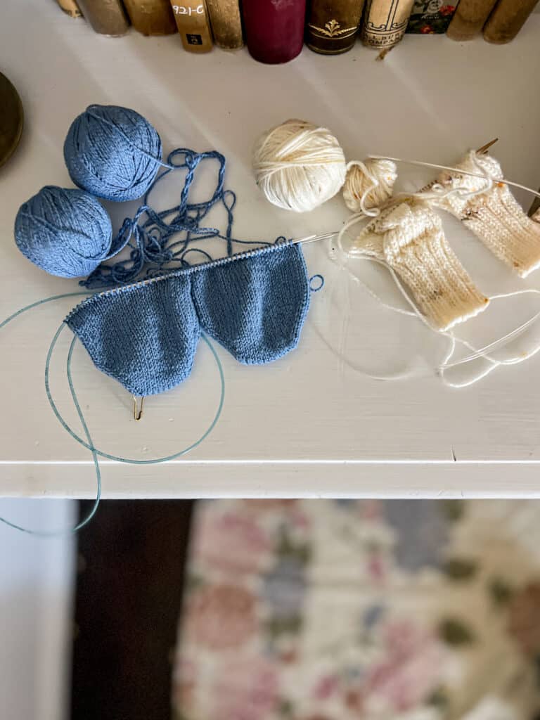 A top-down photo showing two pairs of handknit socks in progress on top of a white dresser surrounded by antique books. One pair of socks is blue, in an adult size, and knit from the toes upward. The other pair of socks is white, in a baby size, and knit from the cuffs downward. Both pairs are being knit two-at-a-time using the Magic Loop method.