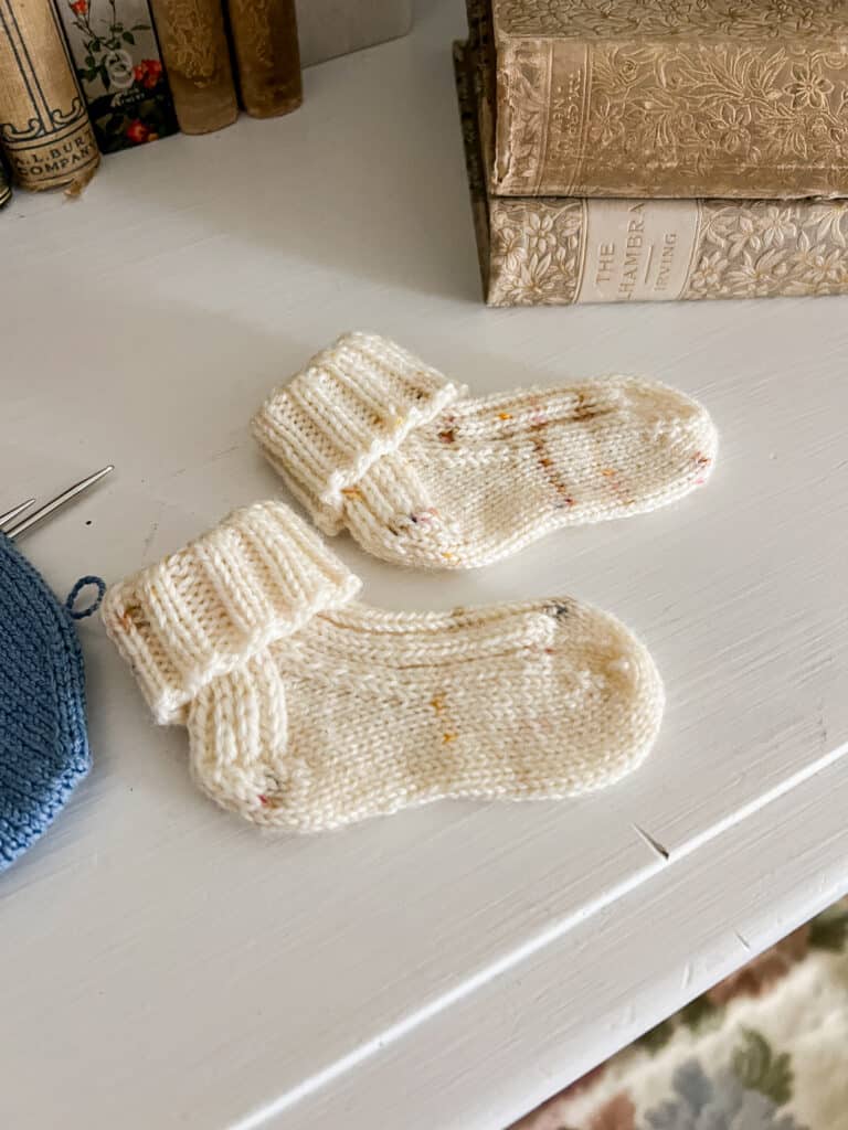 A finished pair of tiny white baby socks with tan speckles. The ribbed cuffs have been folded down.