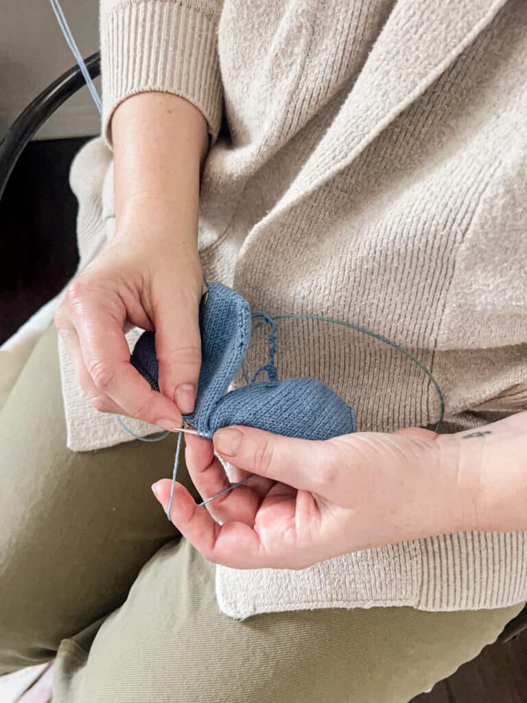 A white woman's hands hold her knitting in her lap as she prepares to make the next stitch. She is working on a pair of blue handknit socks knit two-at-a-time from the toes up using the Magic Loop method.