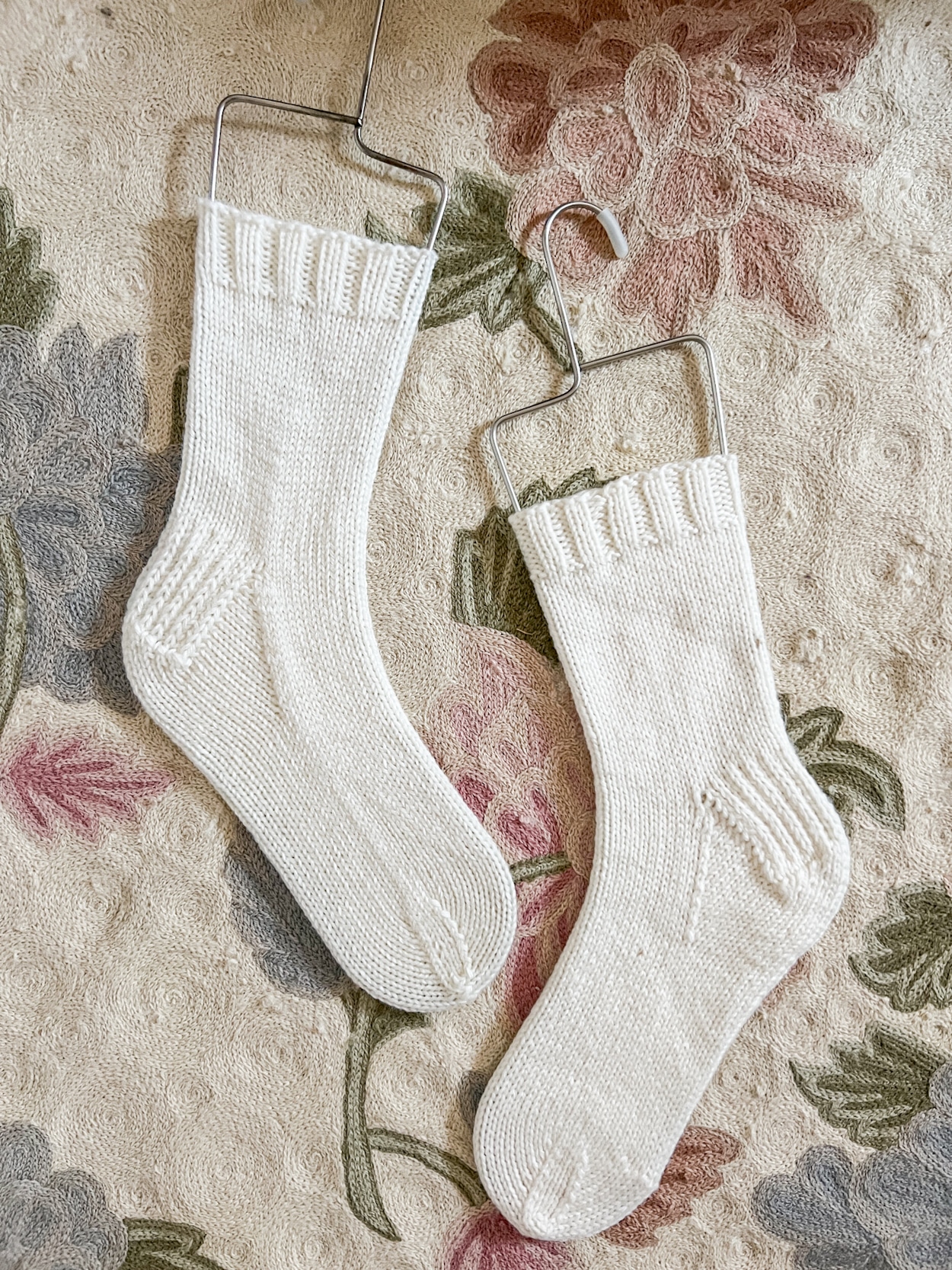 A pair of plain white handknit socks on metal sock blockers rest on a floral rug. The socks are slightly staggered vertically with the toes pointing in opposite directions.