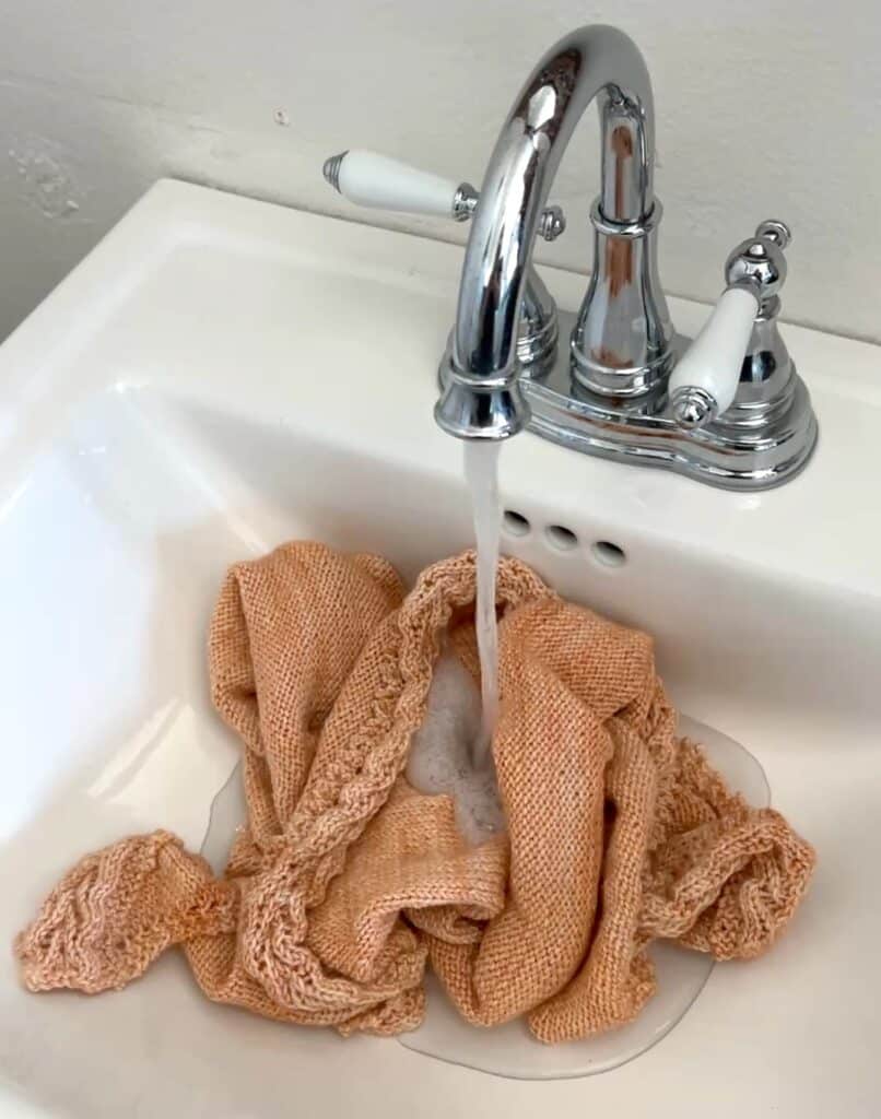 A peach-colored knit shawl soaks in a sink full of cool water, with more water running from the tap into the basin.