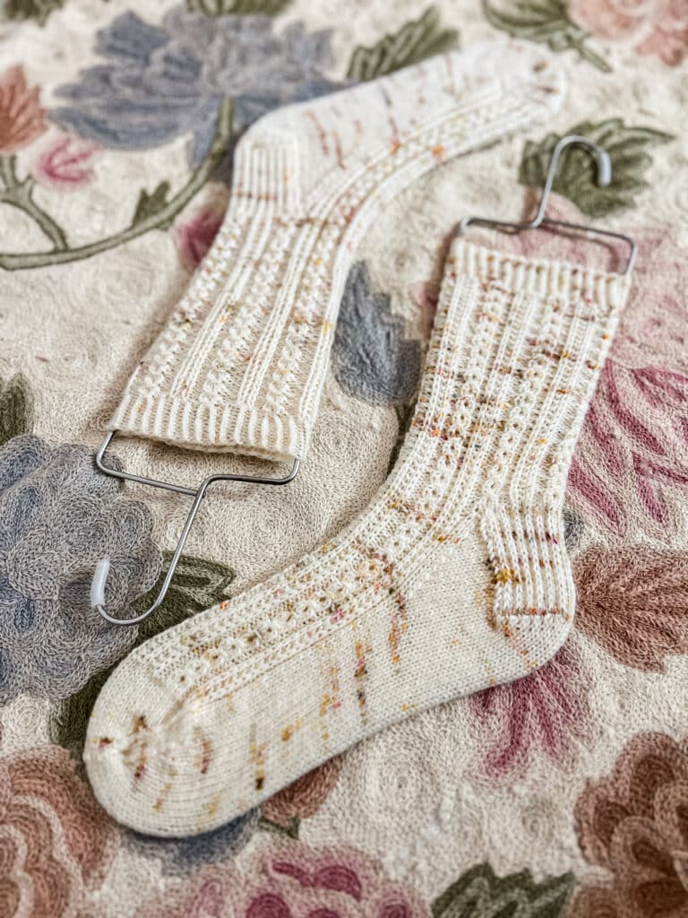 A pair of cream-colored handknit socks with orange and tan speckles dries on a floral rug after a nice soaking in cool water. The socks are stretched on a pair of sock blockers (which, contrary to their name, are not essential to the blocking process).
