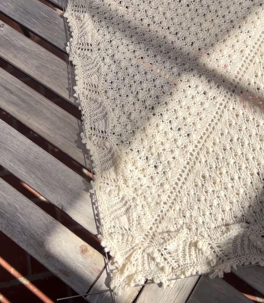 A white shawl dries on a slatted wooden table. The shawl is stretched on blocking wires to ensure a crisp shape.