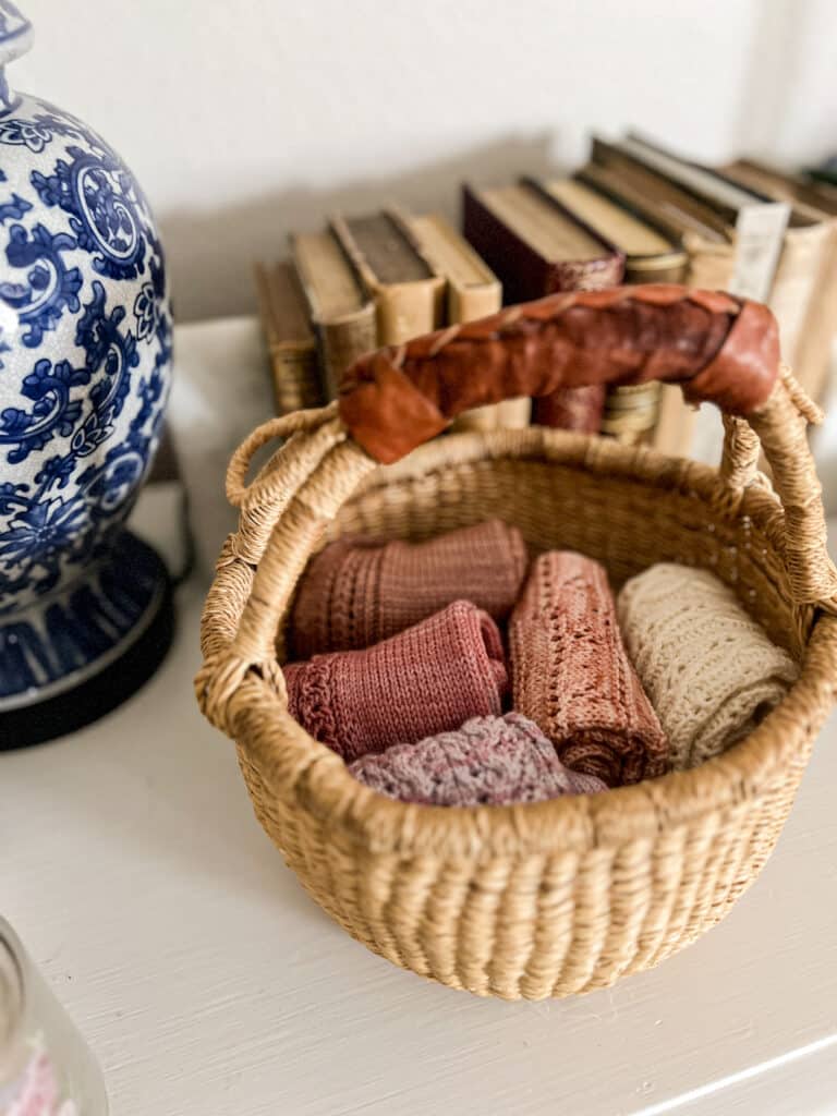 A small woven basket with five pairs of folded pink knit socks in it sits on top of a white dresser surrounded by antique books.