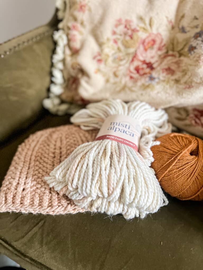 A peach-colored knit hat, a hank of creamy yarn with pastel specks, and a ball of tangerine-colored yarn sit on a green wingback chair in front of a needlepoint pillow with lots of flowers and tassel trim.