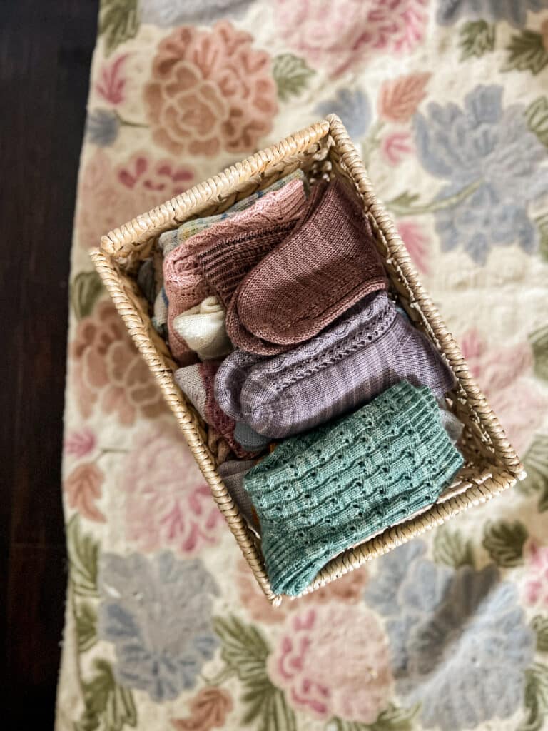 A woven basket filled to the brim with handknit socks sits on a pastel floral rug.
