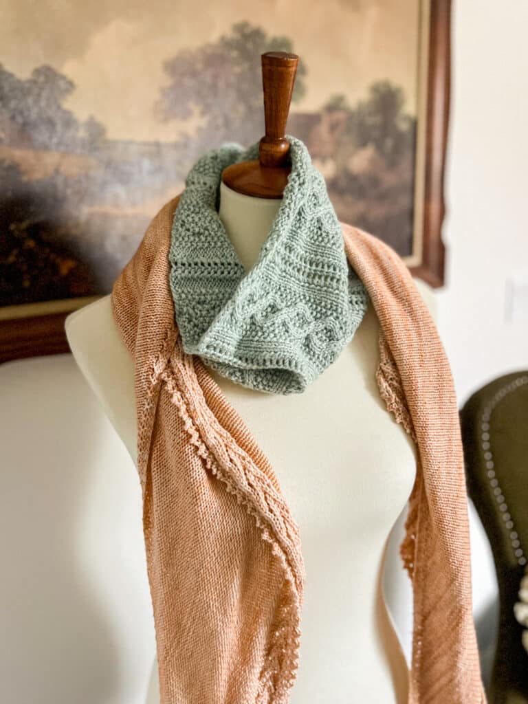 A seafoam green cowl and a peach shawl are draped around the neck of a white dressmaker's form with a wooden topper.