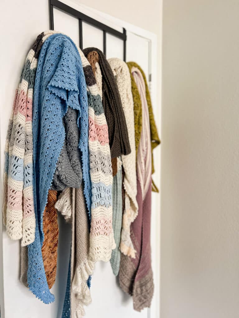 Lots of handknit shawls are hanging from an organizer on the back of a door. The shawls are mostly knit in pastels with the occasional pop of burnt orange and kelp green.