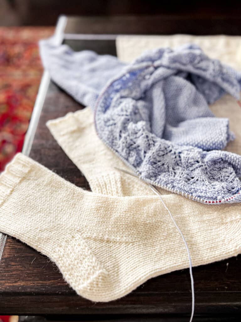 A pair of white handknit socks fills the foreground of this photo. Blurred in the background is a light blue knit shawl in progress on some steel needles.