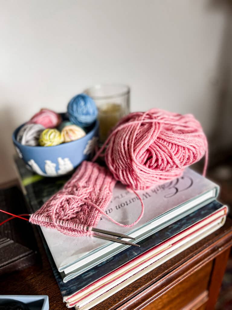 A side-angle photo of a stack of books with a pink knit sock in progress sitting on top of them. There's a pink ball of yarn next to the sock. Blurred in the background is a light blue bowl full of tiny balls of scrap yarn.