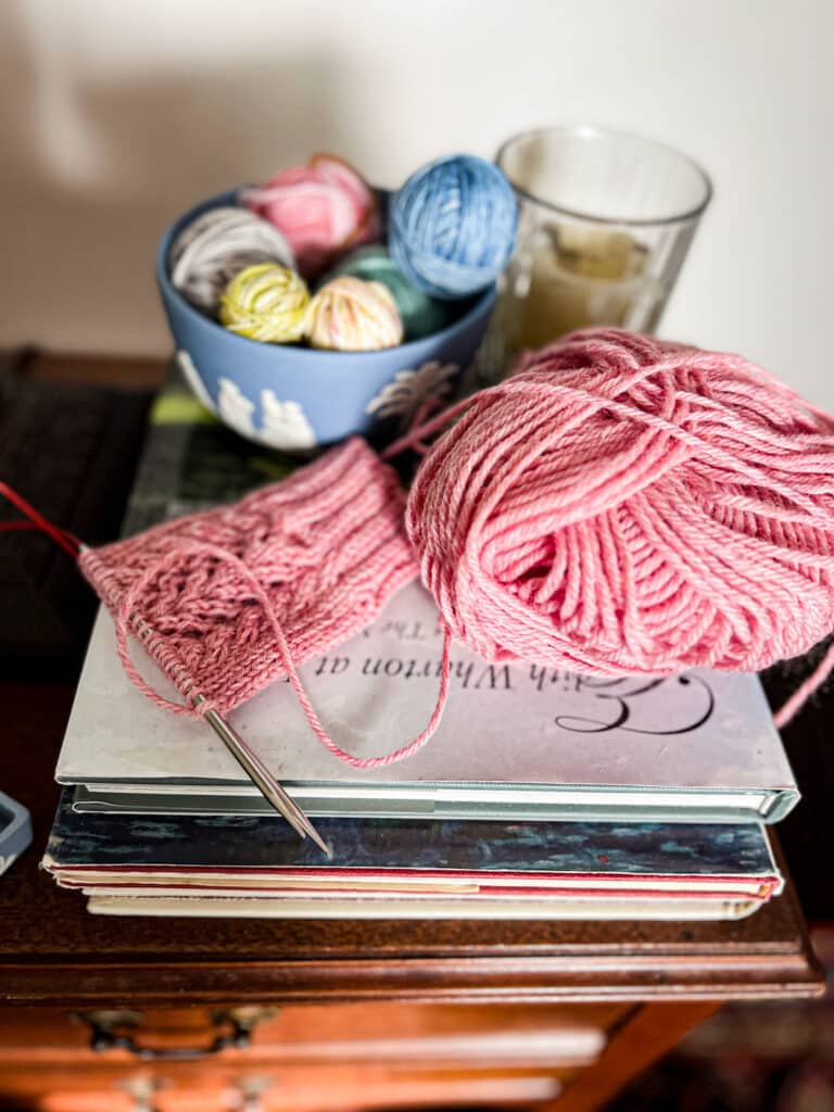 A close up on a pink knit sock in progress sitting on a pile of books. Next to it is a pink ball of yarn, and behind it is a light blue bowl full of lots of tiny balls of scrap yarn.