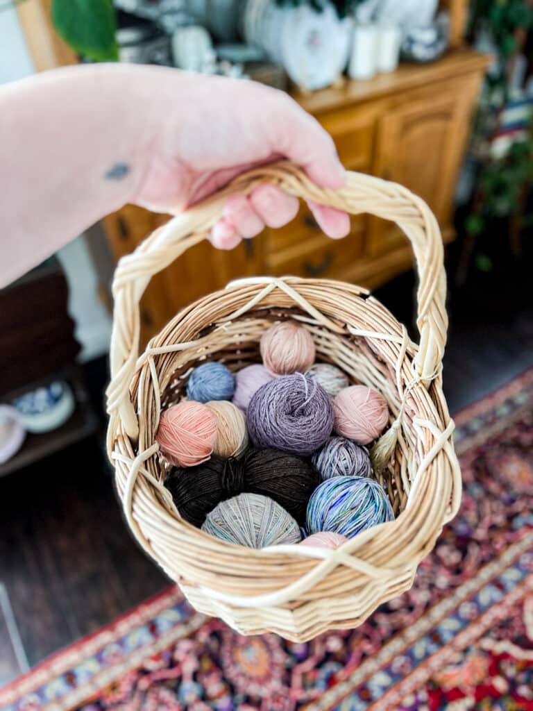A white woman's left hand holds a wicker basket full of balls of leftover yarn in shades of pink, blue, and purple.
