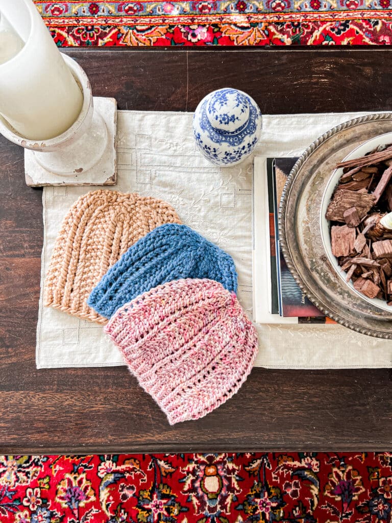 A top-down photo of three hats displayed in a soft arc. All three hats are knit from the same pattern featuring columns of eyelets and seed stitch. The top hat is knit in a clay color, the middle hat is blue, and the bottom hat is pink with speckles. The hats are surrounded by a candle in a ceramic holder, some books, and a bowl full of paperwhite plants.