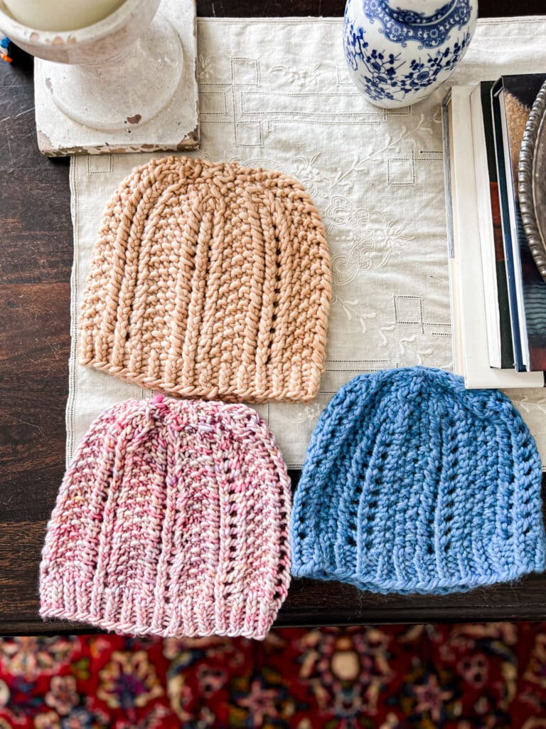 A top-down photo of three hats knit using the same pattern. The upper hat is a clay color, while the bottom two hats are pink with speckles and blue. Each hat features columns of eyelets and seed stitch.