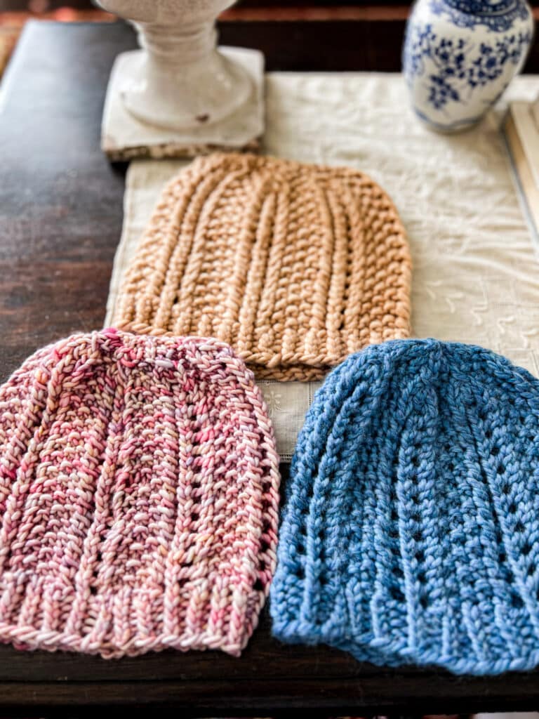 A low-angle photo of three hats knit using the same pattern. The bottom two hats, knit in pink yarn and blue yarn, are in focus, while the upper hat is blurred.