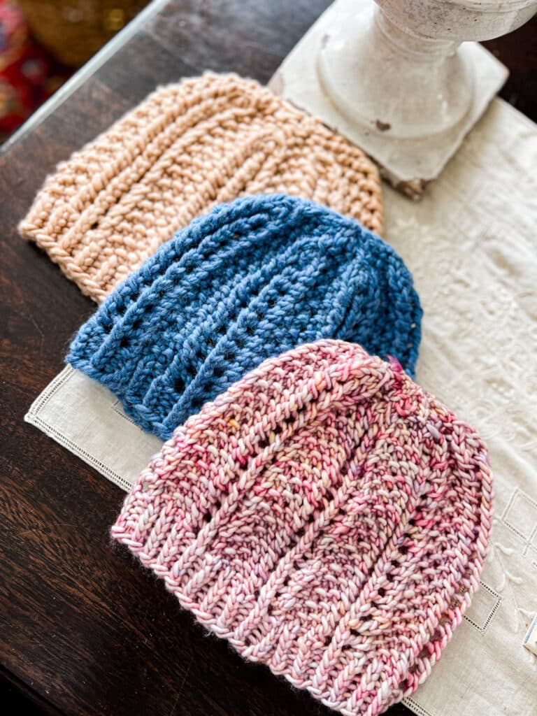 Three handknit hats knit with the same pattern are displayed in a diagonal on a dark wood table. The top left hat is a clay color, the middle hat is blue, and the bottom right hat is pink with speckles. All three feature columns of eyelets and seed stitch.