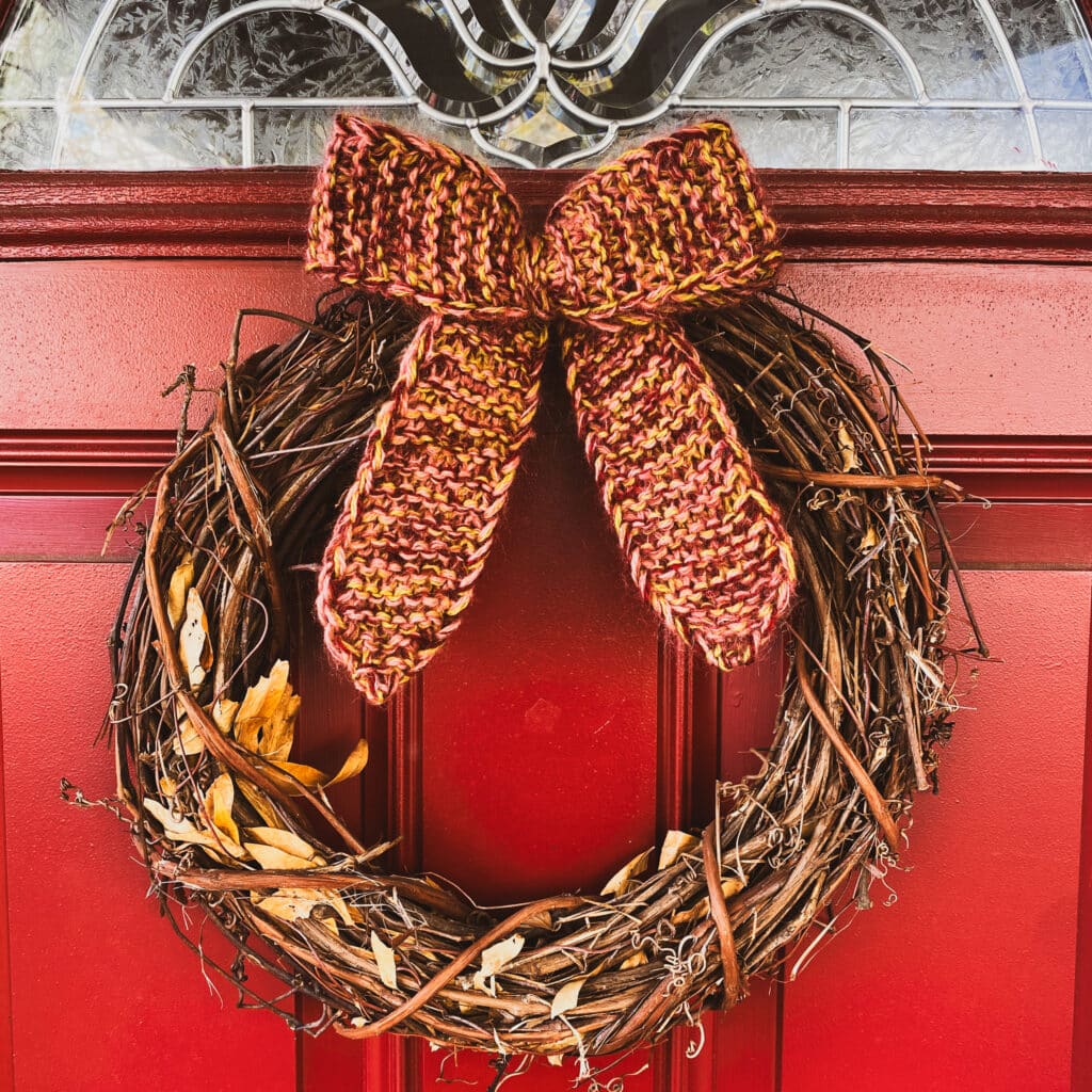 A photo of a brown wreath hanging on a red door. The wreath has been decorated with a knitted bow at the top that was knit in a burgundy yarn with yellow flecks.