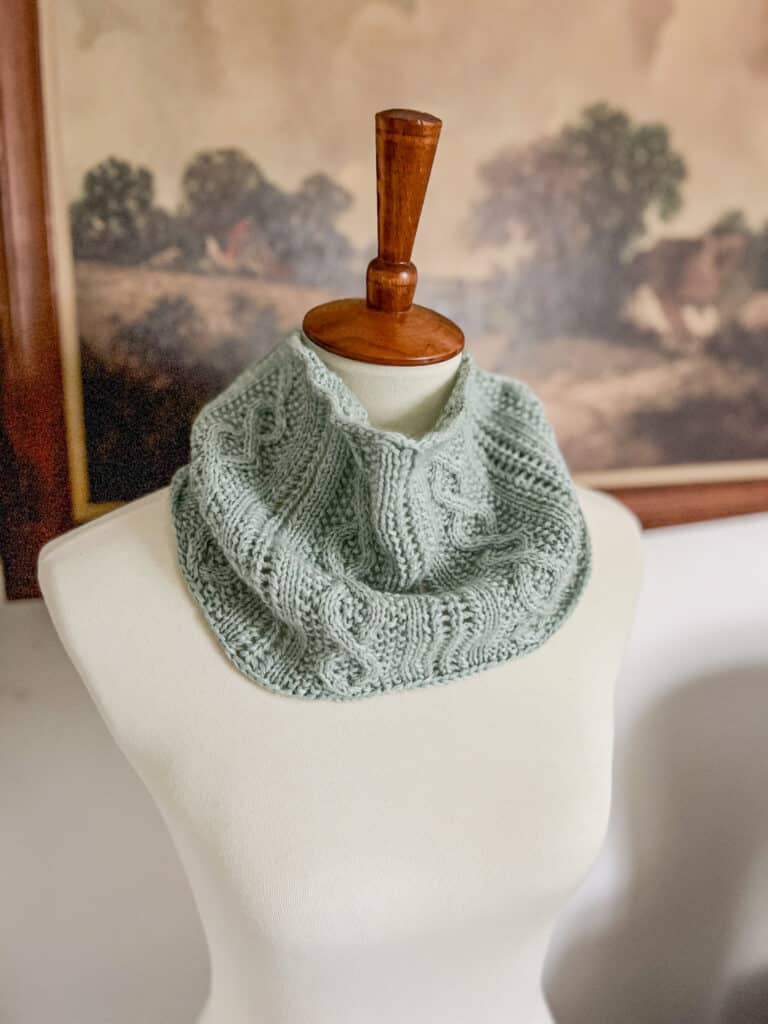 A cabled, handknit cowl in a seafoam green yarn is displayed around the neck of a white dressmaker's form with a wooden top.