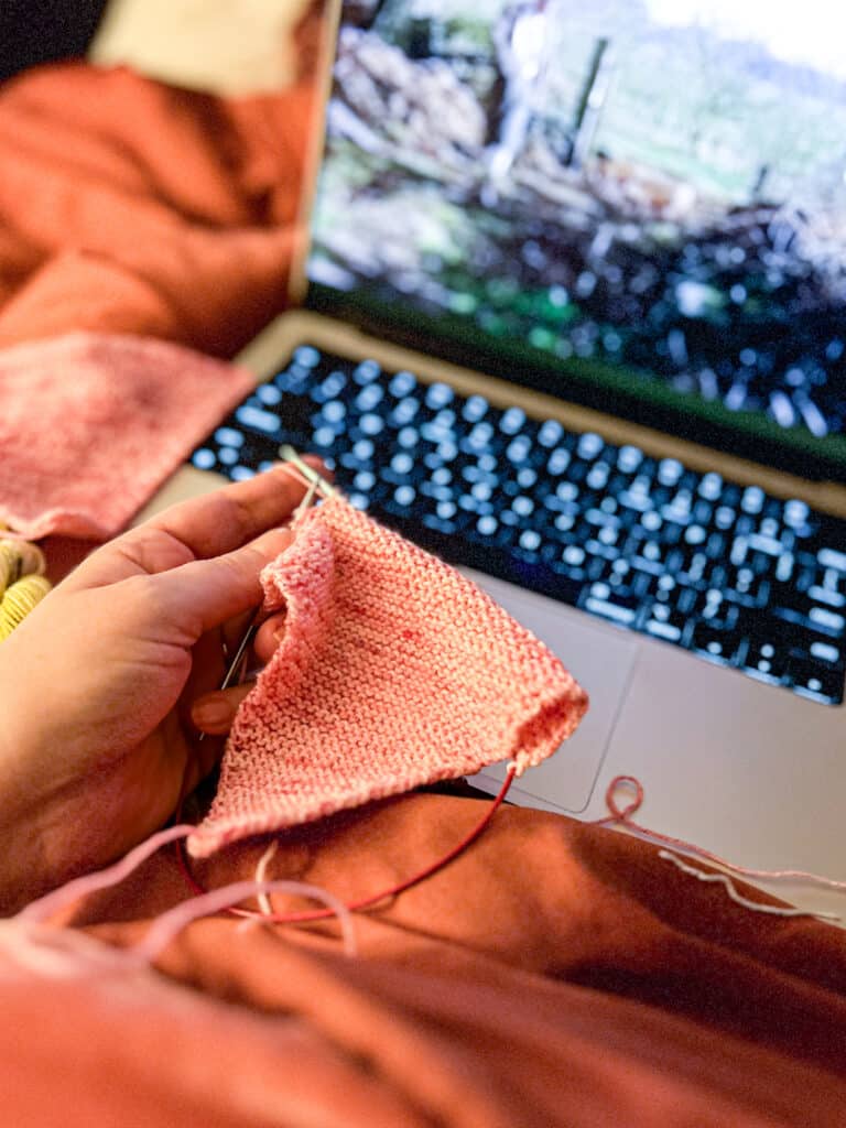 A woman's left hand holds a pink triangle of knitting in front of a blurred laptop screen with a cozy farm scene on it.