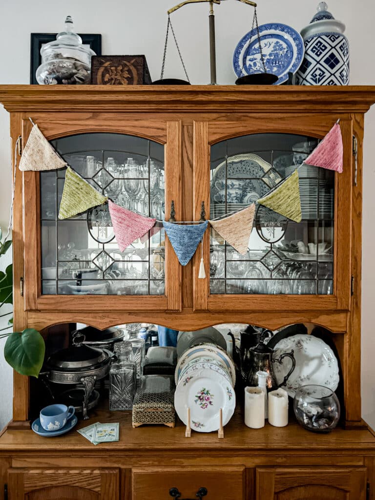 A string of triangle bunting with seven triangles hangs across the front doors of a vintage oak china hutch. The pennants are in shades of pink, blue, yellow, and green.