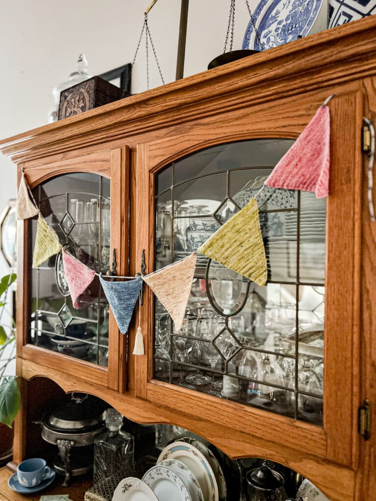 A side-angle photo showing a string of pennant bunting in shades of pink, blue, yellow, and green across the front doors of a vintage oak china hutch.