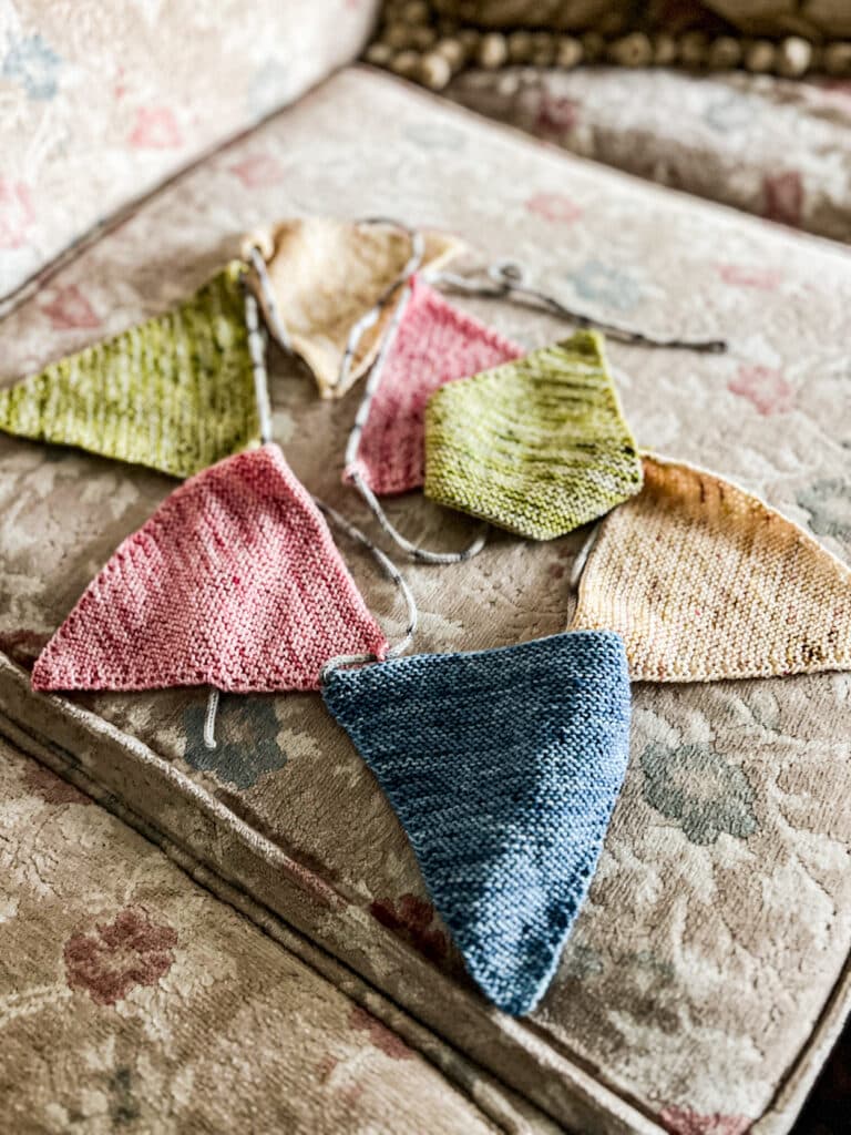 A pile of knit triangles for pennant bunting sits on a vintage sofa cushion with faded floral upholstery.