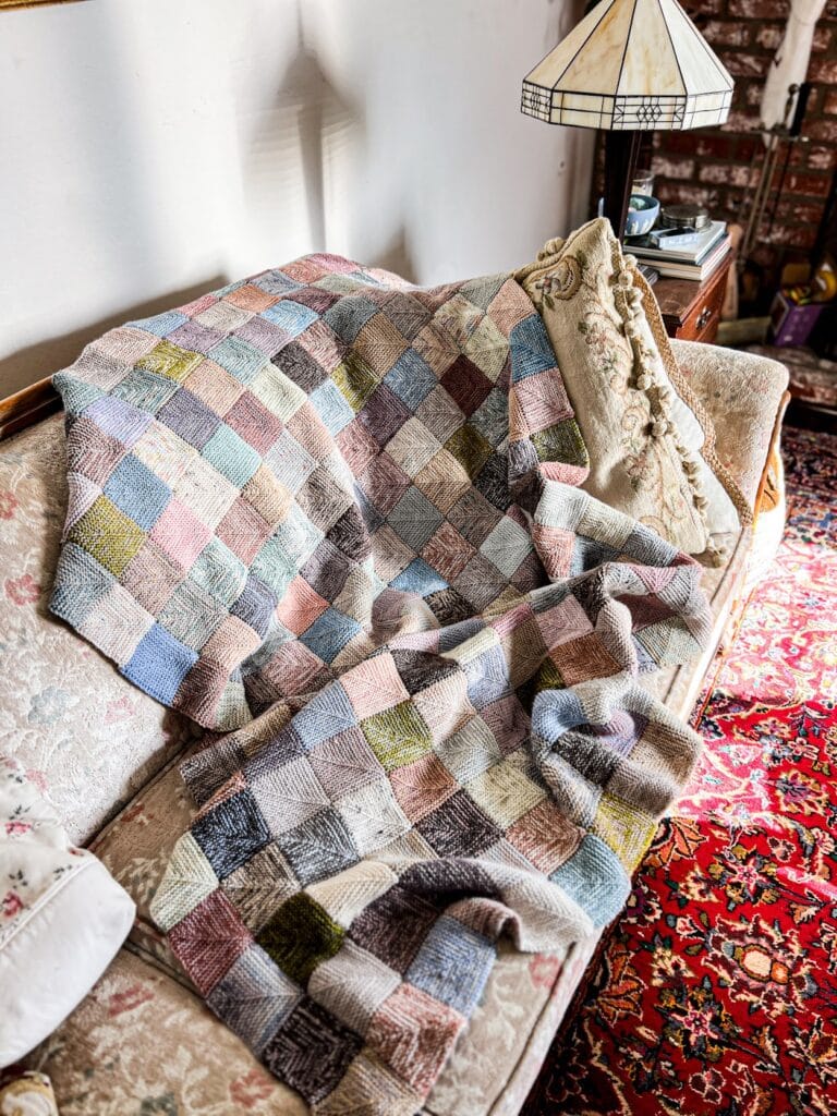 A knit blanket made of multicolored mitered squares is spread messily across a vintage sofa with faded floral upholstery.