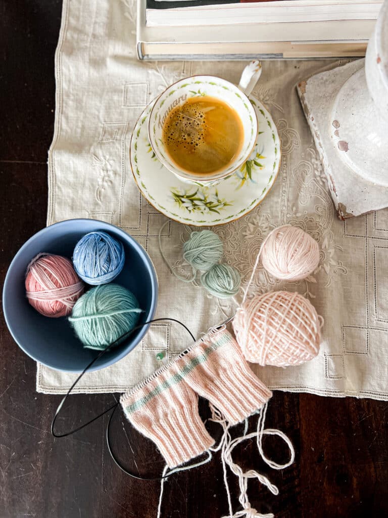 A pair of pink mittens in progress sit on a tabletop surrounded by balls of yarn and a teacup full of very dark coffee. The mittens are being knit two-at-a-time on a set of KnitPro Karbonz needles.