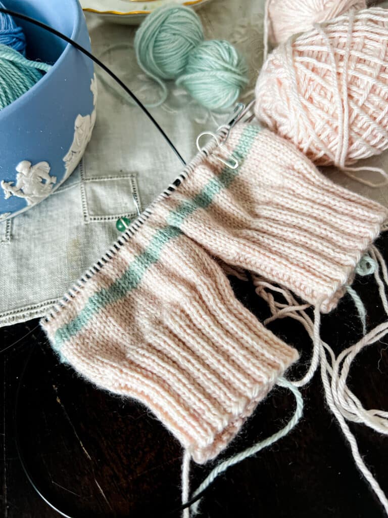 A close-up on a pair of pink mittens in progress on a set of circular needles. The mittens are being knit two-at-a-time on a pair of KnitPro Karbonz needles, and you can see the black carbon fiber peeping between the stitches.