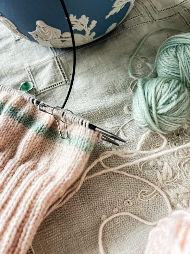 A close-up on the tips of a set of KnitPro Karbonz needles, which are being used to knit a pair of pink mittens. The needles are made of black carbon fiber with metal tips.