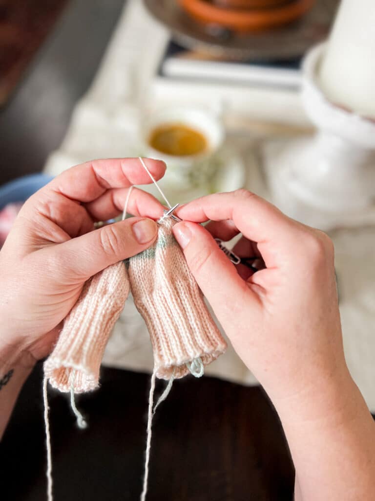 A white woman's hands hold a set of KnitPro Karbons knitting needles while she works on knitting a pair of pink mittens.