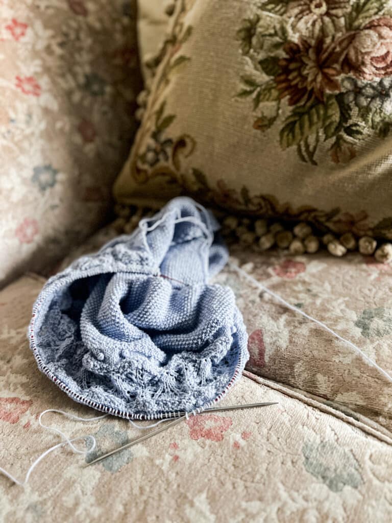 A light blue knit shawl in progress on metal needles sits on the cushion of a vintage sofa with faded floral upholstery. A needlepoint pillow fills the background.