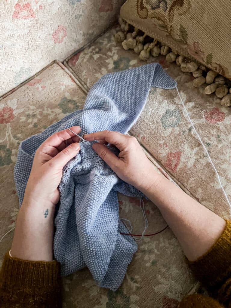 A white woman's hands work on knitting a light blue knit shawl.